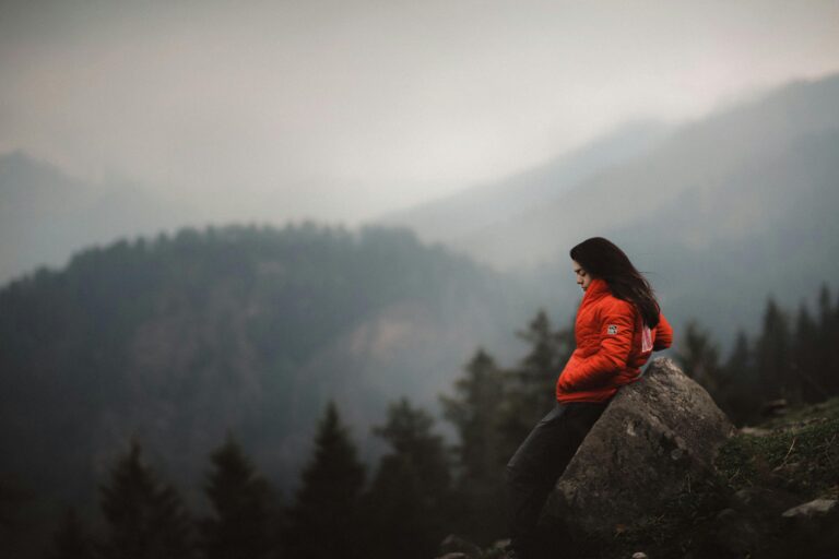 A woman in a red jacket leans against a boulder, enjoying the mountainous view in India.
